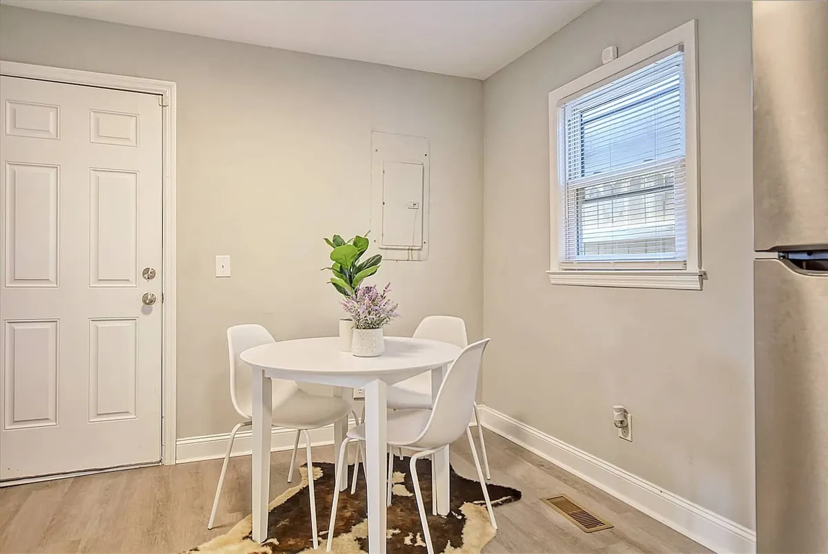 A cozy, casual dining area featuring a round white table with four chairs, a potted plant, and natural light streaming through the window. The space is inviting and perfect for shared meals, morning coffee, or lively conversation.