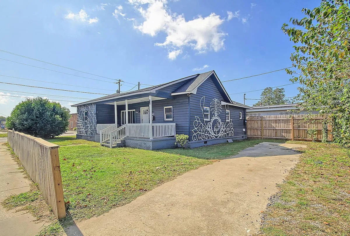 A wider exterior shot with a painted mural on the side of the house, a welcoming porch with steps, a green lawn, and a driveway. The setup suggests space for family gatherings, barbecues, and children playing, all within a quiet, cheerful neighborhood setting.