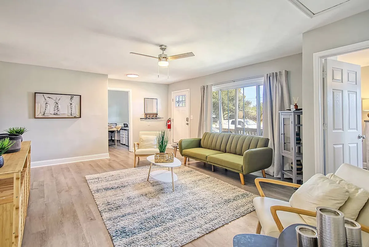 A spacious living room with a green sofa, coffee table, and framed artwork, bathed in natural light from a large window. The layout encourages relaxation and socializing, offering plenty of space for groups to gather before or after their Charleston adventures.