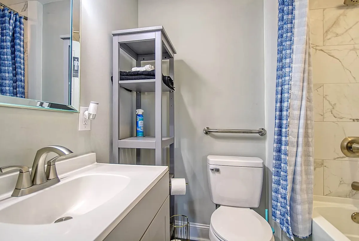 A well-organized bathroom featuring a sink, mirror, toilet, storage shelf, and a shower curtain. The space is tidy and practical, designed for ease in daily routines.