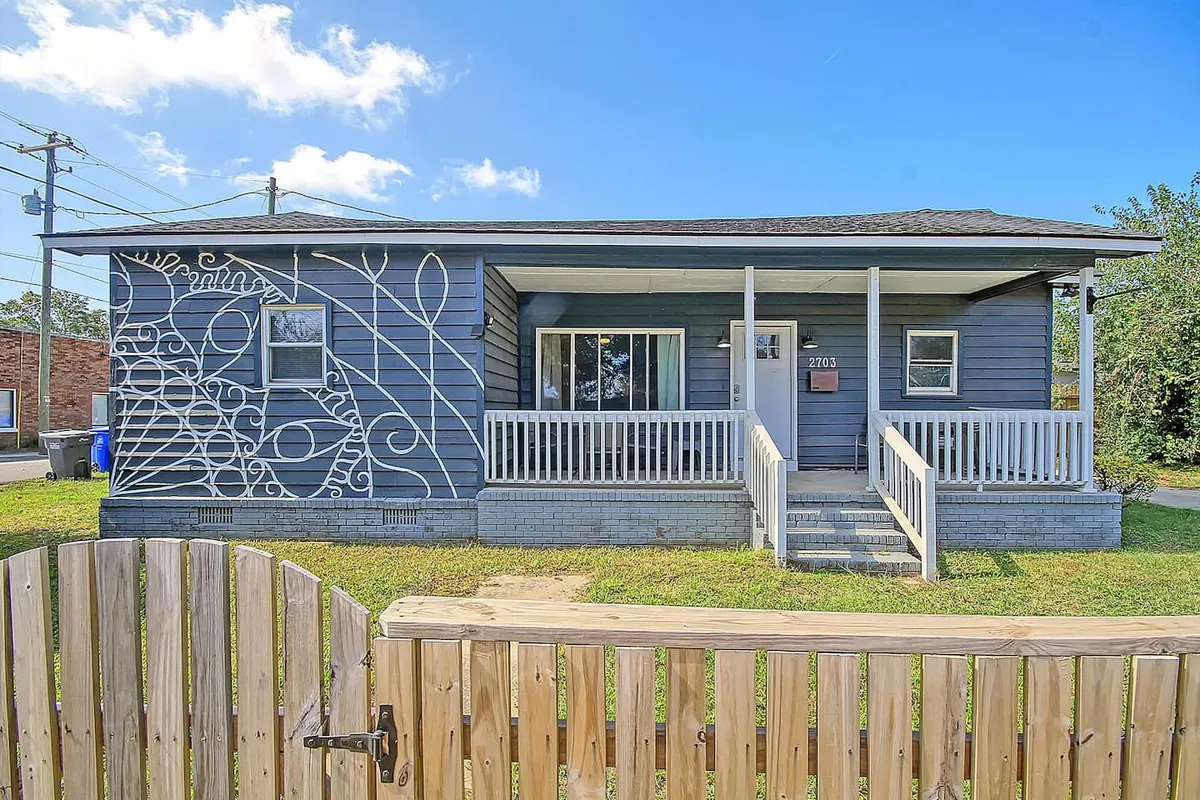 The exterior of a single-story home with a decorative mural on the side, a cozy front porch with steps leading up, and a cheerful atmosphere. People are seen enjoying the porch, engaging with neighbors, and relaxing in a well-kept yard, suggesting a lively, community-oriented environment.