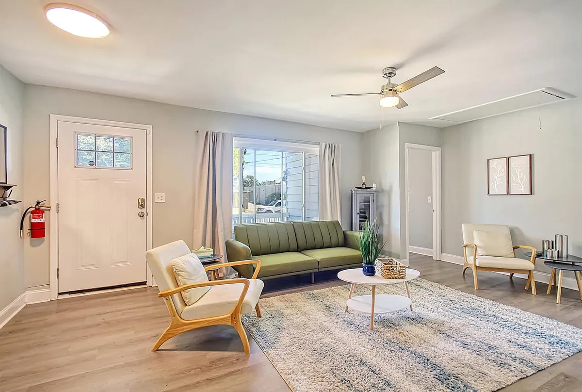 This image features a green sofa as the centerpiece of a sunlit living space, complemented by a white coffee table and light wood chairs. Curtains by the window allow natural light to flood the room, creating a welcoming and communal environment ideal for gatherings.