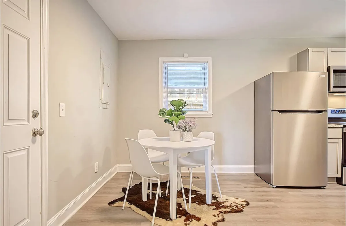 A round dining table surrounded by white chairs and accented with a green plant sits in a cozy, modern dining area. The refrigerator in the background hints at the kitchen's proximity, emphasizing the open-concept flow between spaces.