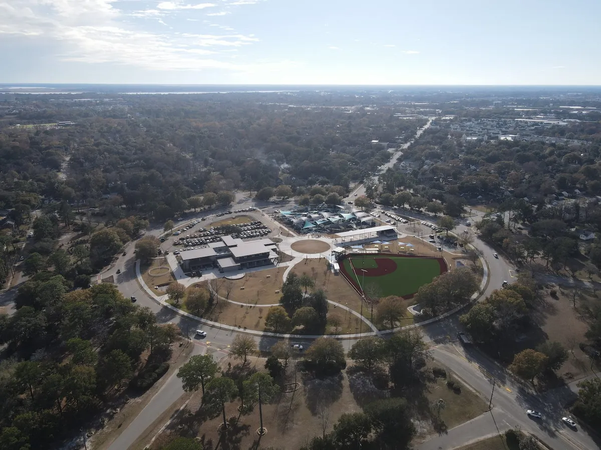 A vibrant community scene with a baseball field, parking area, a community center, and surrounding trees. The image suggests community events, outdoor activities, and a lively neighborhood environment.