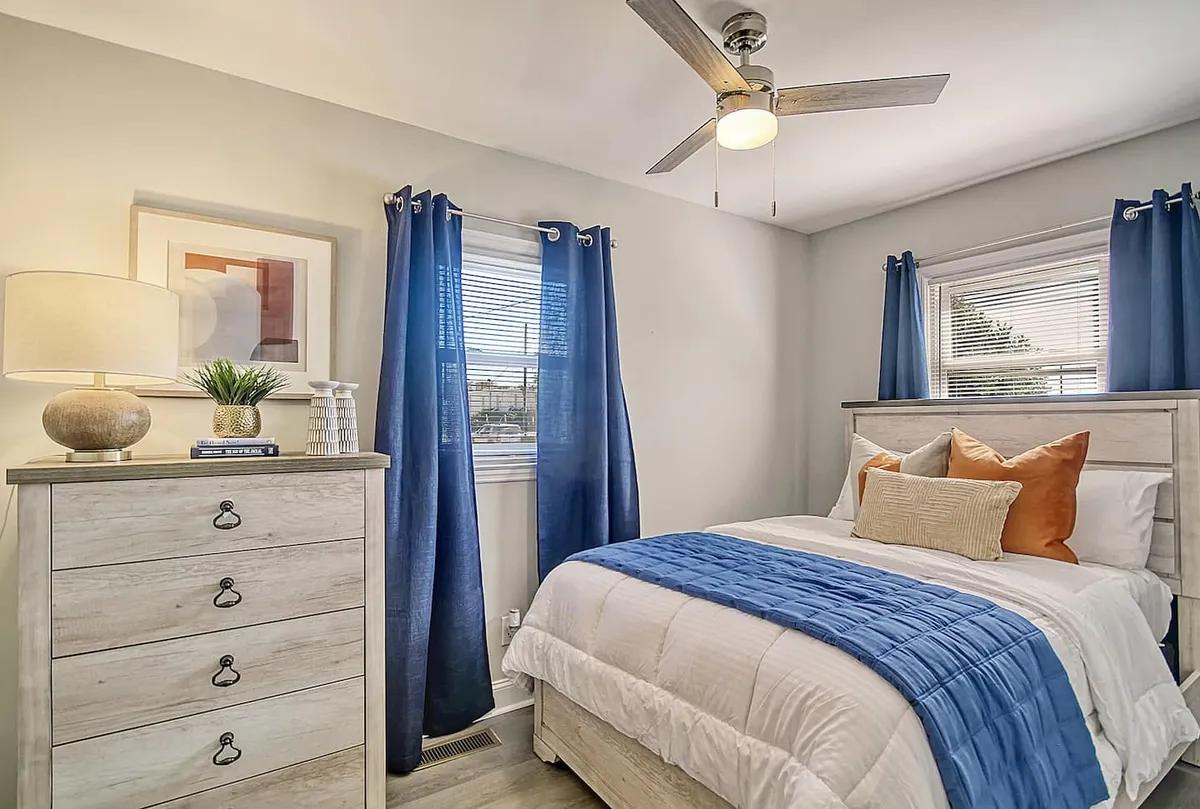 This image features a cozy bedroom with blue and white bedding, a dresser, blue curtains, and a bedside lamp. Natural sunlight streams through the windows, creating a serene and calming atmosphere ideal for rest and relaxation.