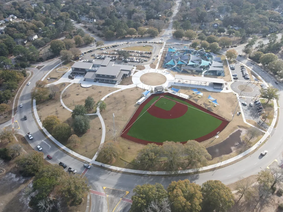 A lively exterior scene featuring a baseball field, a bustling community center, a playground in the distance, and a parking lot full of activity. Families gather for games and picnics, with children playing and adults socializing, all set against a backdrop of green spaces and community infrastructure.