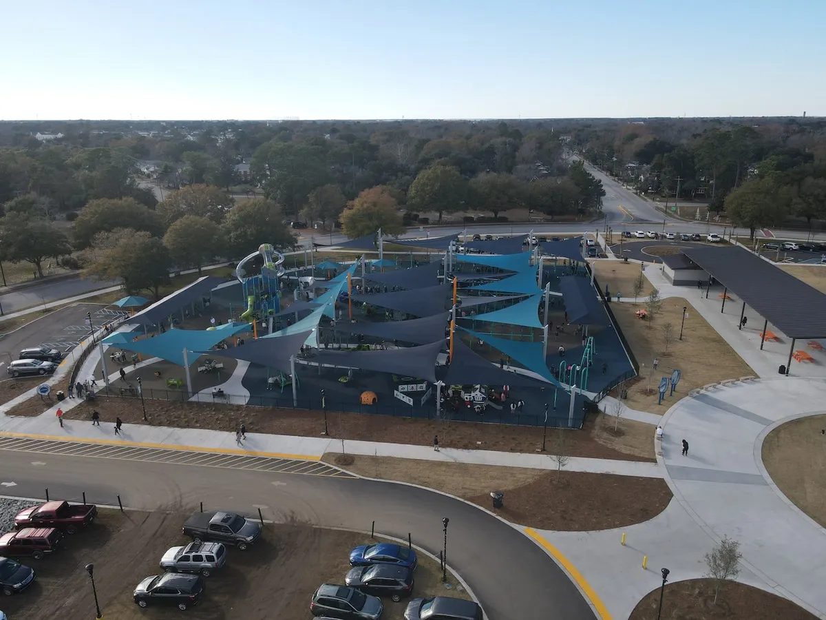 A vibrant playground scene with modern play equipment, colorful shade structures, winding pathways, and families gathered in the background. Children can be seen playing energetically, while adults relax nearby, and there are vehicles parked in an adjacent lot, suggesting easy access for visitors.