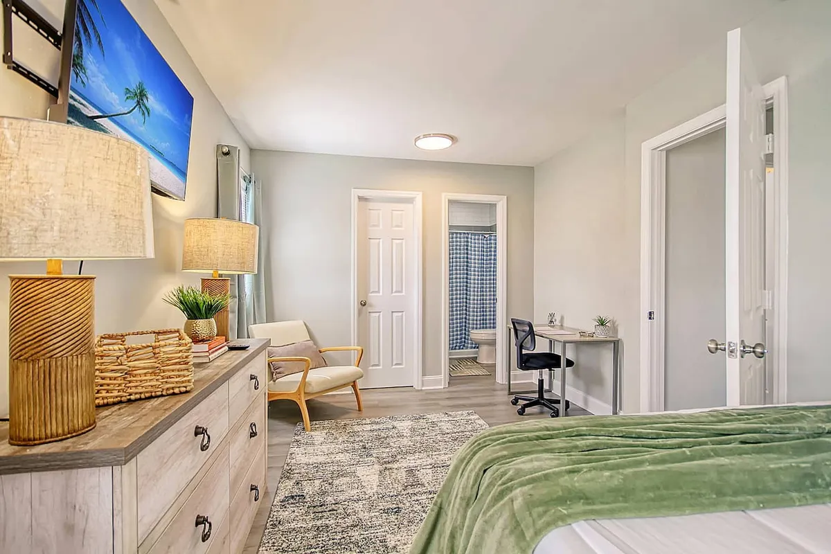 A tranquil bedroom with a bed dressed in a green throw blanket, a wooden dresser, two lamps, and a piece of beach-themed artwork. The space has a calm, cozy vibe, offering a peaceful morning retreat or a comfortable space for reading or remote work.