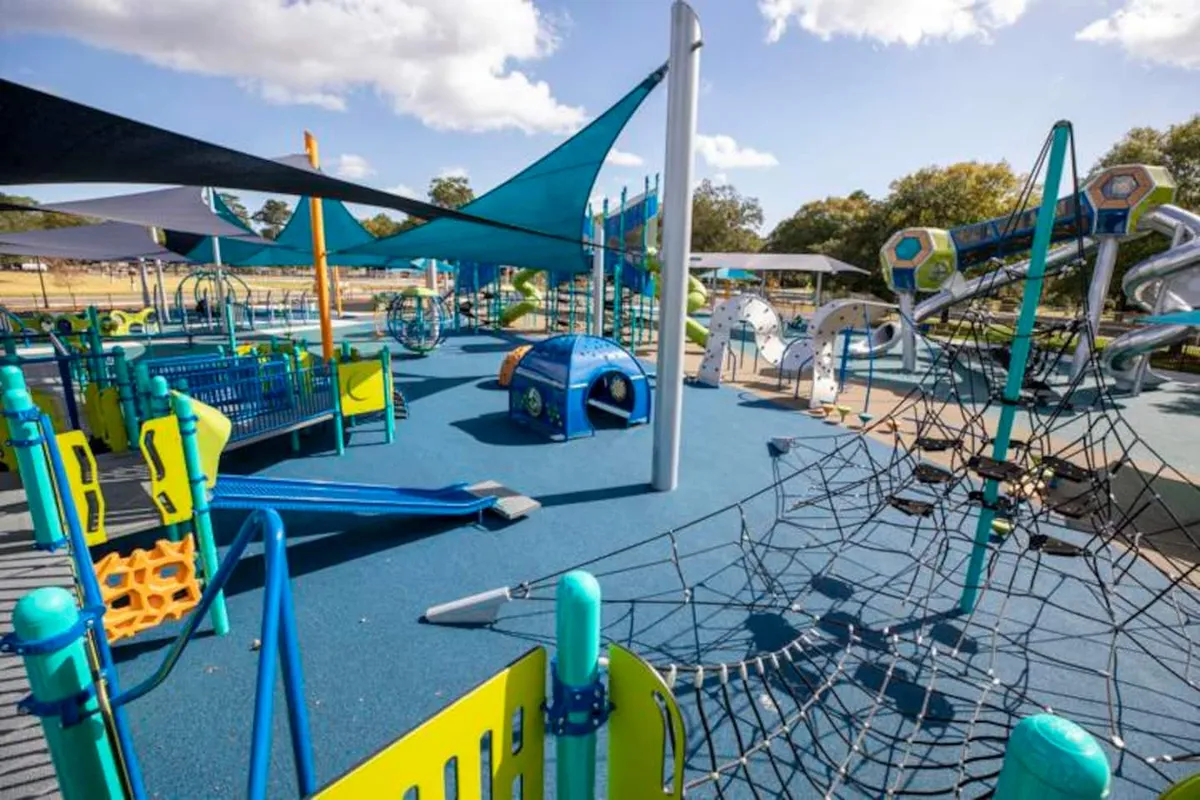 A vibrant playground scene with colorful play structures, slides, climbing nets, and shade sails. Children are actively playing, while families gather and picnic around the playground. The setting is joyful and energetic, representative of nearby amenities for families and urban explorers alike.